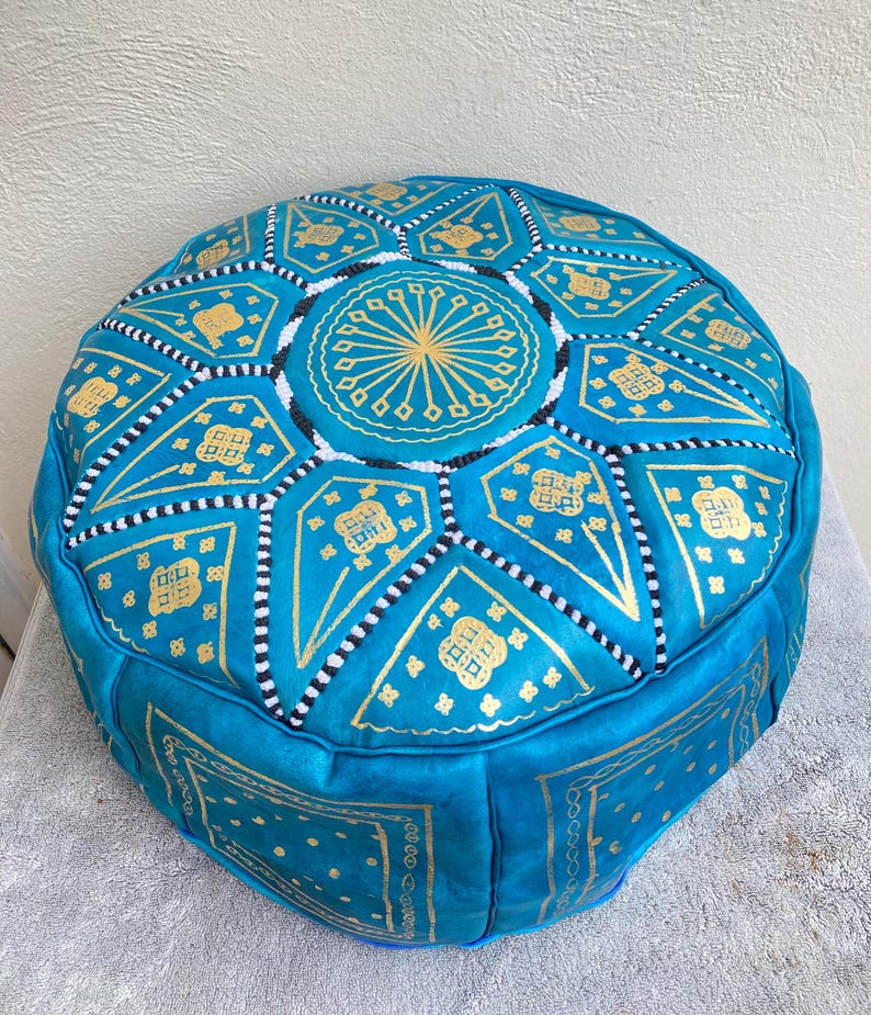 May include: A turquoise blue leather pouf with gold and white geometric patterns. The pouf is round and has a tufted top.
