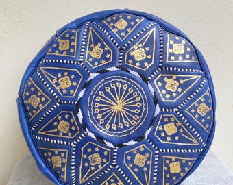 May include: A blue leather pouf with a gold geometric pattern. The pouf is round and has a flat top.