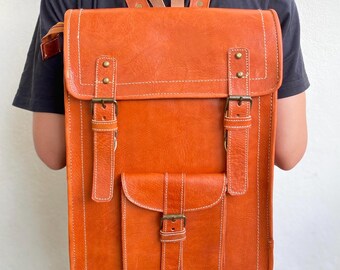 May include: A brown leather backpack with two large front pockets and a smaller pocket with a buckle closure. The backpack has a top flap with two buckles and a stitched design.