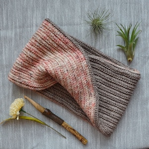 May include: A crochet cowl in a two-toned color scheme of brown and pink. The cowl is made with a textured stitch and is shown on a gray surface with a crochet hook and air plants.
