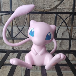 May include: Pink and white Pokemon figurine of a Mewtwo with blue eyes. The figurine is sitting on a black metal grate.
