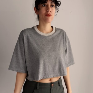 May include: A cropped grey t-shirt with a cream-coloured neckline and short sleeves. The shirt is paired with dark green trousers. The model is wearing a flower hair clip.