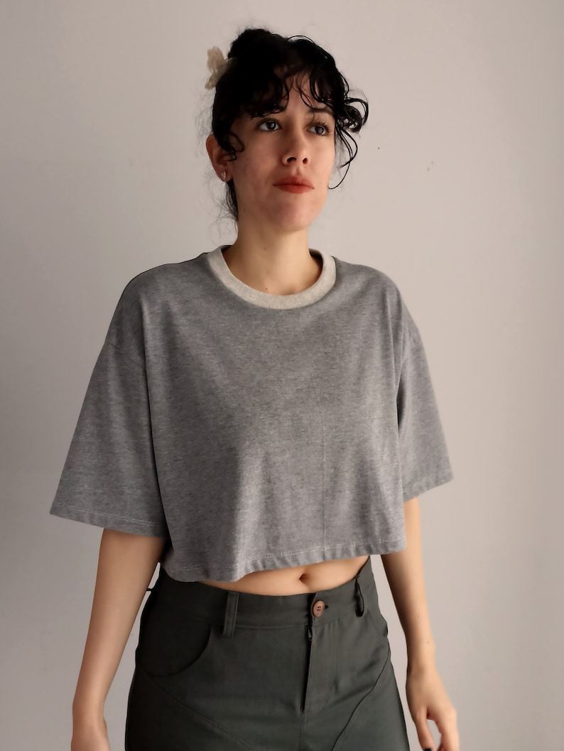 May include: A cropped grey t-shirt with a cream-coloured neckline and short sleeves. The shirt is paired with dark green trousers. The model is wearing a flower hair clip.