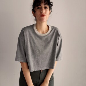 May include: A grey cropped t-shirt with a white neckline. The shirt has short sleeves and a relaxed fit. The model is wearing dark green trousers.