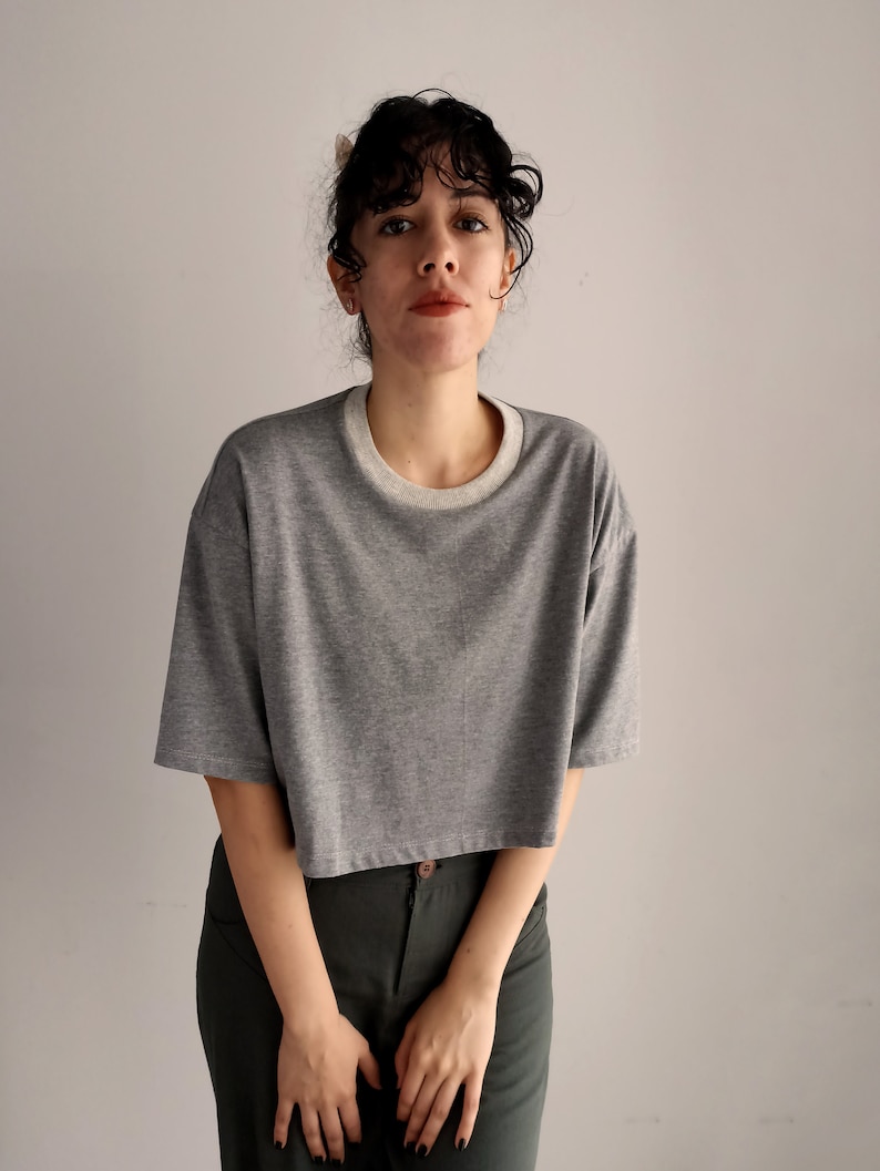 May include: A grey cropped t-shirt with a white neckline. The shirt has short sleeves and a relaxed fit. The model is wearing dark green trousers.
