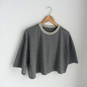 May include: A grey crop top with short sleeves and a light grey neckline, suspended from a wooden hanger. The top has a loose fit and a minimalist design, suitable for casual wear. The shirt is made of a soft material.