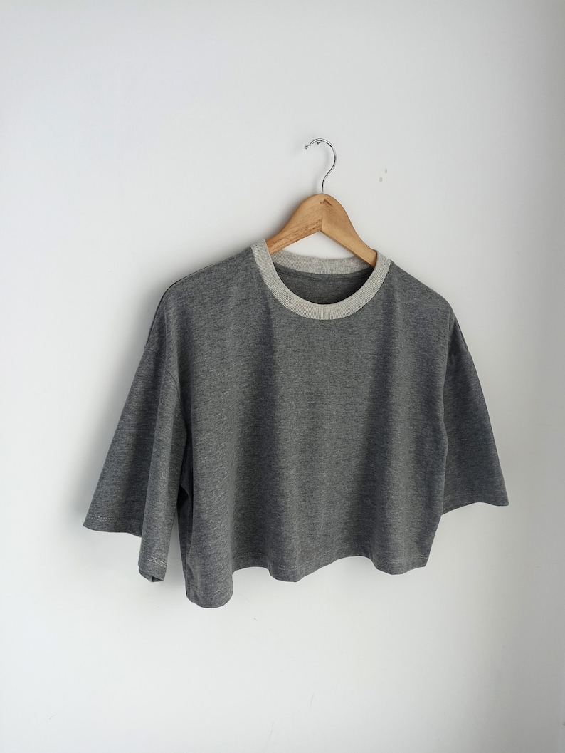 May include: A grey crop top with short sleeves and a light grey neckline, suspended from a wooden hanger. The top has a loose fit and a minimalist design, suitable for casual wear. The shirt is made of a soft material.