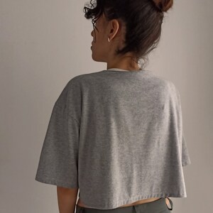 May include: A grey, cropped t-shirt with short sleeves. The shirt is made of a soft material and has a relaxed fit. The model is wearing the shirt with green trousers.