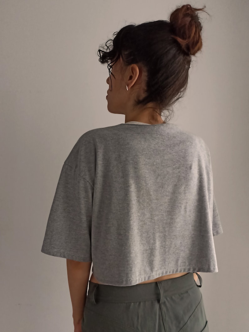 May include: A grey, cropped t-shirt with short sleeves. The shirt is made of a soft material and has a relaxed fit. The model is wearing the shirt with green trousers.