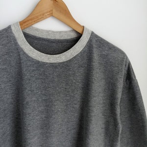 May include: A grey short-sleeved top with a light grey crew neck collar. The shirt is suspended on a wooden hanger against a white backdrop. The fabric looks to be a soft, knitted material.