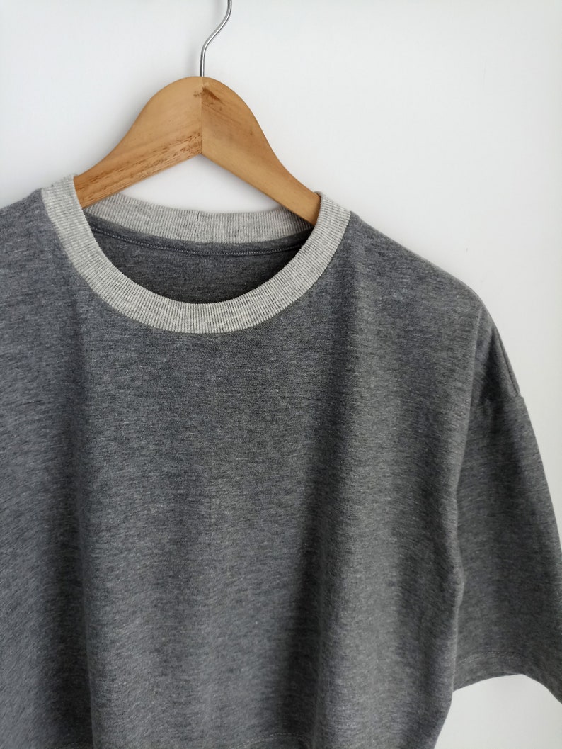 May include: A grey short-sleeved top with a light grey crew neck collar. The shirt is suspended on a wooden hanger against a white backdrop. The fabric looks to be a soft, knitted material.