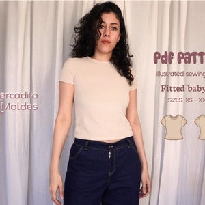 Puede incluir: Una mujer con una camiseta de bebé beige ajustada y vaqueros azul oscuro. La imagen incluye el texto "Pdf PATTERN, illustrated sewing guide, Fitted baby tee, SIZES: XS - XXXL" y el logo "Mercadito Moldes".