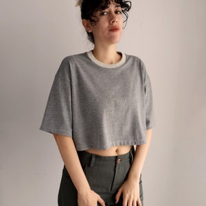 May include: A grey cropped t-shirt with a light grey neckline and short sleeves. The shirt is paired with dark green trousers. The model is wearing a hair accessory.