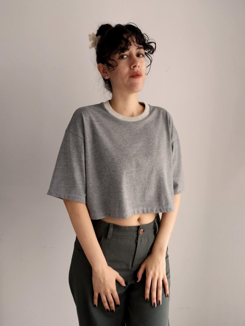 May include: A grey cropped t-shirt with a light grey neckline and short sleeves. The shirt is paired with dark green trousers. The model is wearing a hair accessory.