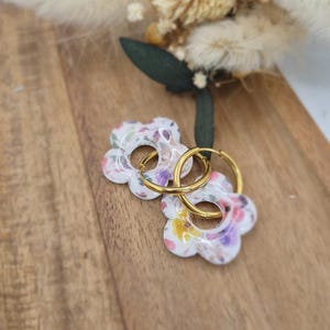 May include: A pair of gold hoop earrings featuring white flower-shaped charms with a colourful floral pattern. The charms have a smooth, glossy finish and are attached to thin, circular gold-toned metal hoops that pass through the centre of each flower.