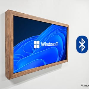 May include: A walnut-framed screen displaying the Windows 11 logo and abstract blue background. The screen is mounted on a white wall. A Bluetooth symbol is visible on the right. The Yaeger Woodworks logo is in the lower left.