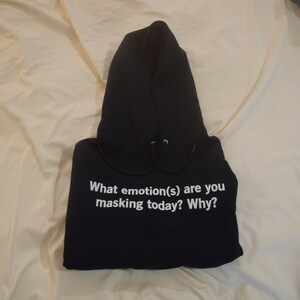 Może przedstawiać: Czarna bluza z kapturem z nadrukiem "What emotion(s) are you masking today? Why?" w kolorze białym na przodzie.