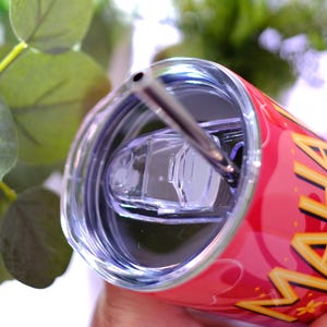 May include: A red insulated tumbler with a silver straw and a clear lid. The tumbler has the word "MAHA" in yellow and gold lettering. The background includes green foliage.