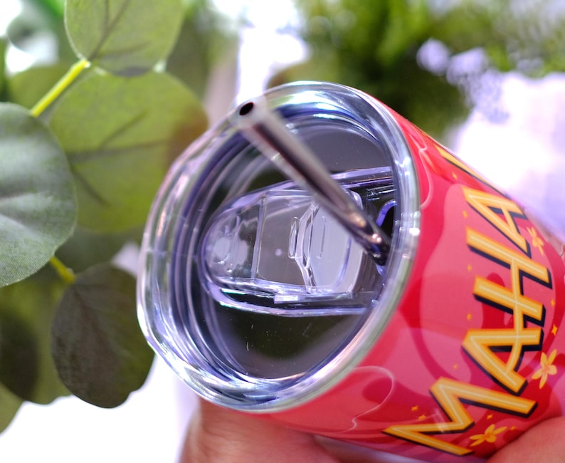 May include: A red insulated tumbler with a silver straw and a clear lid. The tumbler has the word "MAHA" in yellow and gold lettering. The background includes green foliage.