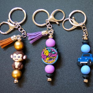 May include: Three keychains with silver-tone hardware. One features a monkey charm, gold beads, and a brown tassel. Another has a purple tassel, a round charm with text, and purple beads. The third has a blue jeep charm and blue beads.