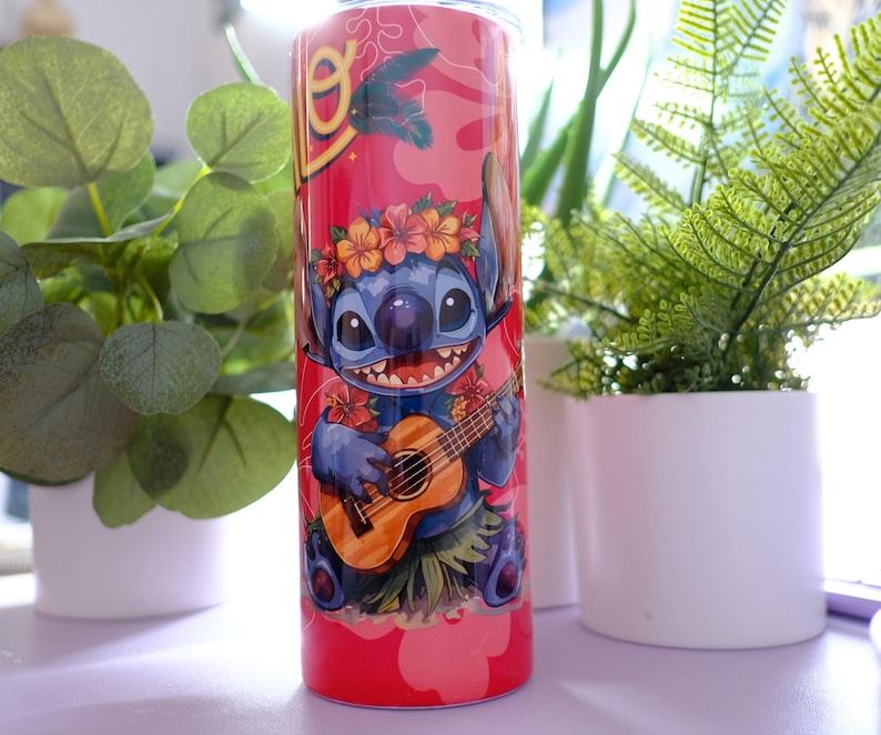 May include: A red tumbler featuring a cartoon character playing a ukulele, adorned with a floral lei. The design includes a tropical theme with a pink background and palm tree accents. The tumbler is cylindrical and appears to be made of stainless steel.