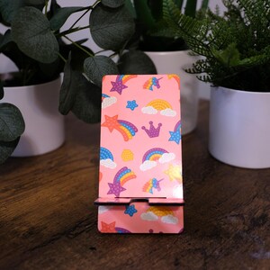 May include: A pink phone stand with a colorful pattern of rainbows, stars, clouds, and a crown. The stand is made of wood and has a slot to hold a phone. The stand is decorated with a repeating pattern of unicorns.