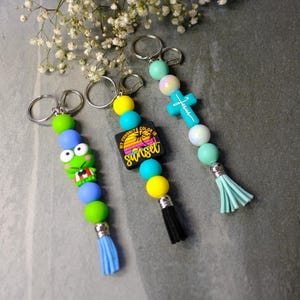 May include: Three beaded keychains with silver rings. One features a green frog, another has a black square with a sunset graphic and the text "My Favorite Color is Sunset", and the last has a blue cross with the word "Jesus". Each has a tassel.