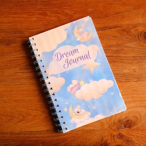 May include: A spiral-bound notebook with a light blue sky background featuring white clouds, yellow stars, and illustrations of small animals resting on crescent moons. The cover displays the text Dream Journal in purple script lettering.