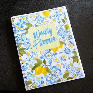 May include: A spiral-bound weekly planner featuring a white background with a blue and yellow tile-inspired pattern, green leaves, and yellow lemons. A central light yellow rectangular label displays the text Weekly Planner in blue script.