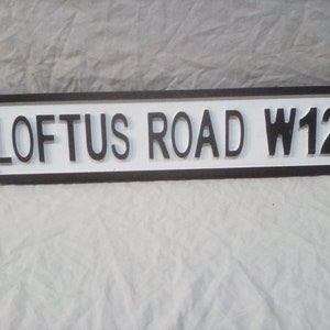 Könnte beinhalten: Ein schwarz-weißes Straßenschild mit dem Text "LOFTUS ROAD W12" in erhabenen Buchstaben.