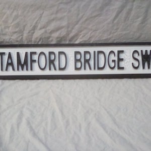 May include: A rectangular street sign with the text "STAMFORD BRIDGE SW6" in black, raised lettering. The sign has a white background and a black border with decorative rounded corners. The sign is likely made of metal.