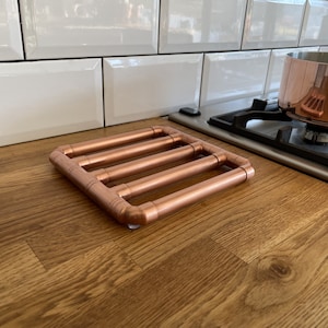 May include: A copper-colored metal trivet with a square design, featuring four parallel bars. The trivet is made of copper pipes and is designed to protect surfaces from hot cookware. A copper pot sits on a gas stove in the background.