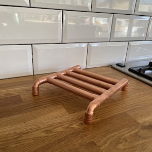 May include: Copper pipe trivet with a rectangular design. The trivet has four horizontal bars and four curved legs. The copper has a shiny, metallic finish. The trivet is on a wooden surface.