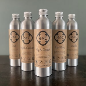 May include: Five silver metal bottles with screw-on caps are shown. Each bottle has a brown label with the logo "R&C" and a scent name: Oud, English Orchard, Fig & Cassis, Earl Grey, and Vanilla. The bottles are for diffuser refills.