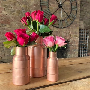 Pode incluir: Três vasos de cobre de tamanhos variados, cada um com rosas frescas. As rosas são de um vermelho e rosa vibrantes. Os vasos são cilíndricos com faixas horizontais, colocados sobre uma mesa de madeira. O fundo apresenta uma parede de tijolos e um relógio grande.