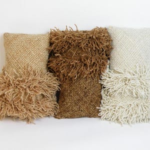 May include: Three rectangular throw pillows arranged side by side. The left pillow is light tan, the center is dark brown, and the right is off-white. Each pillow features a woven texture on the top half and a thick, shaggy fringe layer.