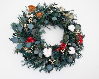 May include: A circular wreath composed of various dried flowers and greenery. The wreath features dark green foliage, blue-green eucalyptus leaves, white cotton bolls, and pops of red and orange flowers. The wreath is set against a white background.