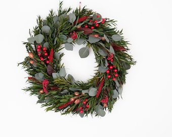 May include: A circular wreath composed of green foliage, accented with red berries and dried flowers. The wreath is set against a white background, showcasing its natural colors and textures. A decorative piece for seasonal home decor.