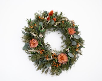 May include: A circular wreath featuring a variety of dried foliage and flowers. The wreath includes green leaves, orange flowers, and small white accents. The wreath is set against a white background, creating a natural, rustic aesthetic.