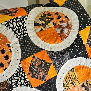 May include: A black and orange Halloween quilt with a variety of patterns, including pumpkins, bats, and spiders. The quilt is made with a variety of fabrics and is quilted with a white thread. The quilt is folded and displayed on a chair.