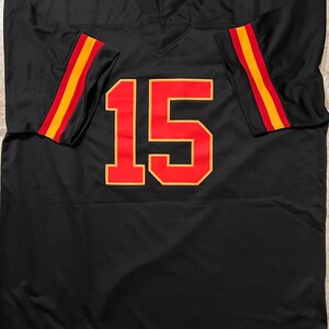 Puede incluir: Camiseta de f&uacute;tbol negra con el n&uacute;mero 15 en rojo y dorado. Las mangas tienen rayas rojas y doradas. La camiseta tiene cuello en V.