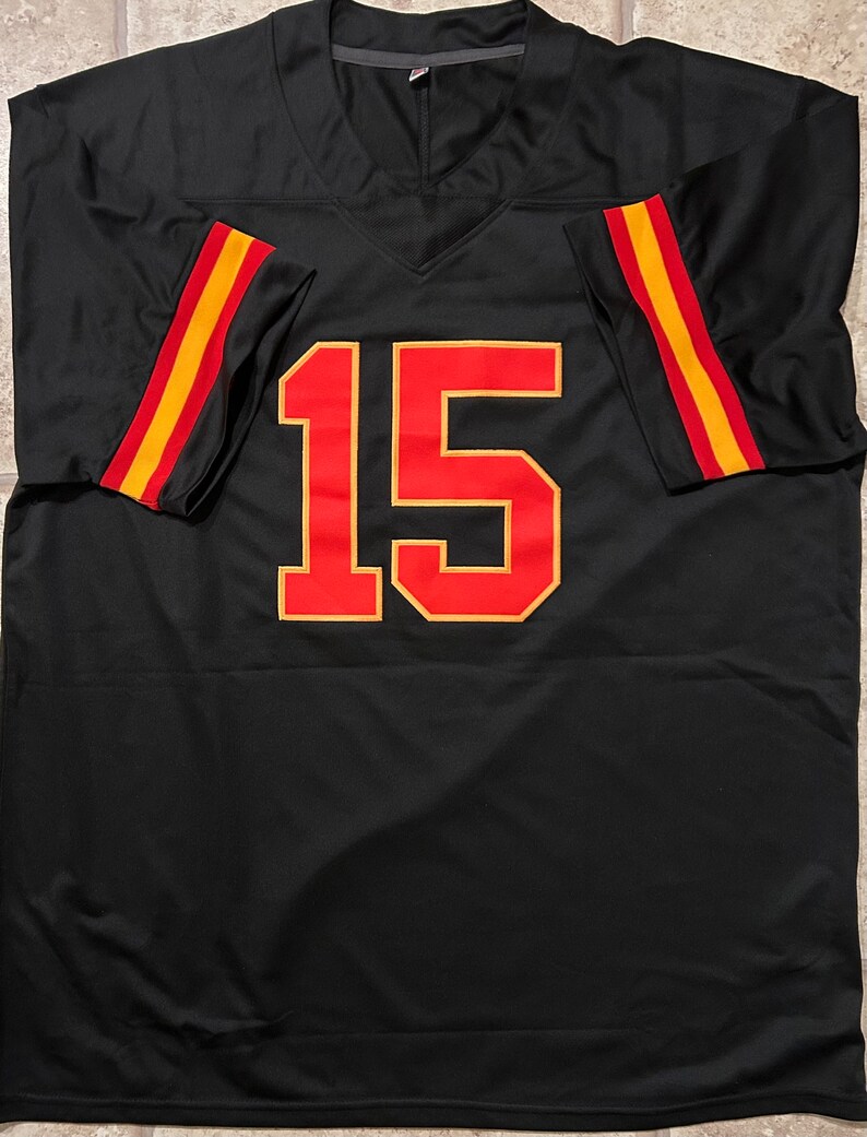 Puede incluir: Camiseta de f&uacute;tbol negra con el n&uacute;mero 15 en rojo y dorado. Las mangas tienen rayas rojas y doradas. La camiseta tiene cuello en V.