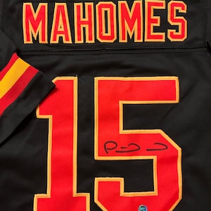 Puede incluir: Camiseta de f&uacute;tbol negra con el nombre "MAHOMES" en letras rojas y doradas en la parte superior de la espalda. El n&uacute;mero "15" se muestra en rojo con ribete dorado en la parte delantera. La camiseta tiene una firma con tinta negra.