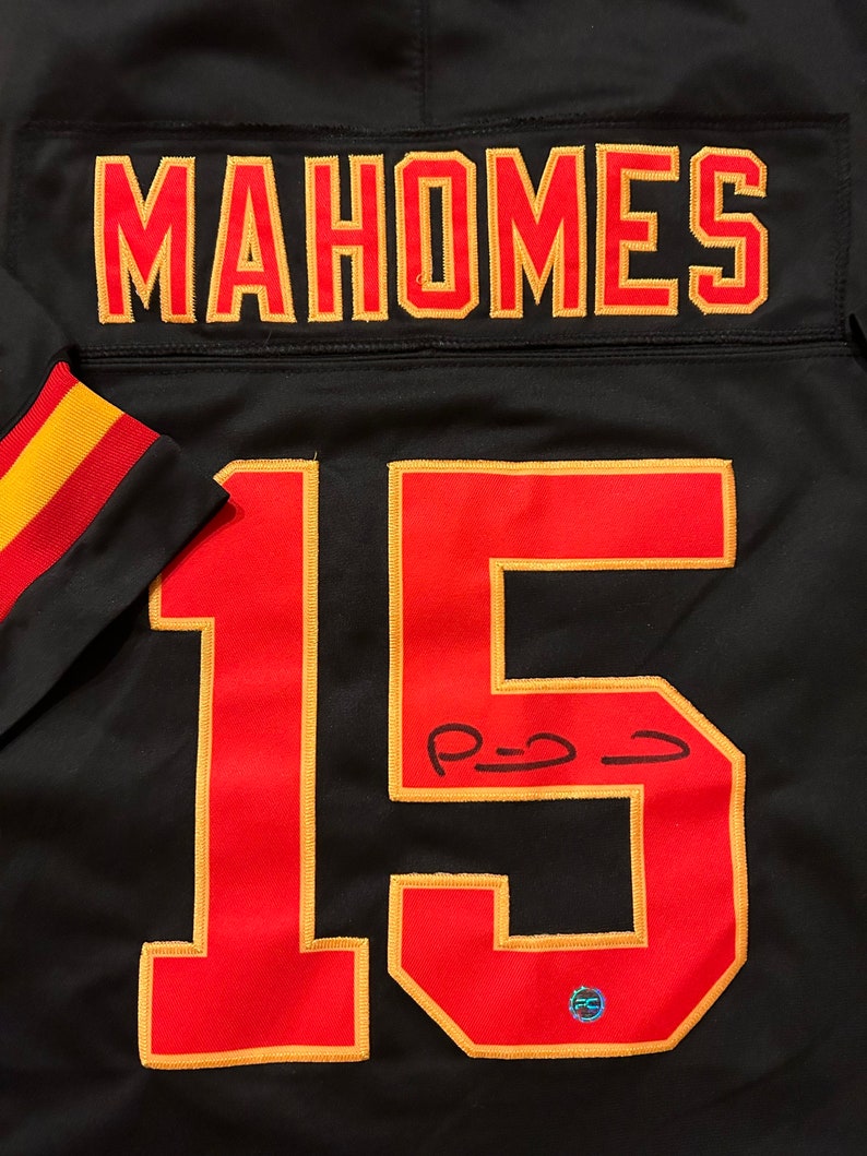 Puede incluir: Camiseta de f&uacute;tbol negra con el nombre "MAHOMES" en letras rojas y doradas en la parte superior de la espalda. El n&uacute;mero "15" se muestra en rojo con ribete dorado en la parte delantera. La camiseta tiene una firma con tinta negra.