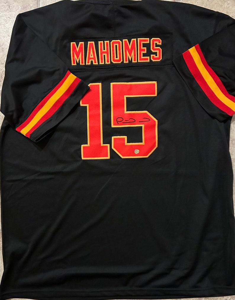 Puede incluir: Camiseta negra con el nombre "MAHOMES" en letras rojas y doradas en los hombros. El n&uacute;mero "15" es rojo con un contorno dorado en la parte delantera. Las mangas tienen rayas rojas y doradas.