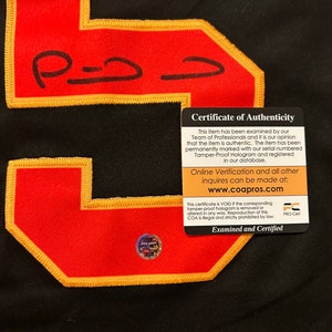 Puede incluir: Primer plano de una camiseta negra con un gran n&uacute;mero 5 rojo con bordes dorados. El n&uacute;mero tiene una firma en tinta negra. Se adjunta un certificado de autenticidad, con informaci&oacute;n sobre la autenticidad del art&iacute;culo.