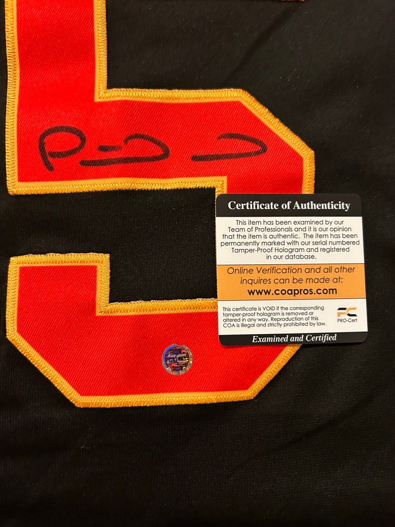 Puede incluir: Primer plano de una camiseta negra con un gran n&uacute;mero 5 rojo con bordes dorados. El n&uacute;mero tiene una firma en tinta negra. Se adjunta un certificado de autenticidad, con informaci&oacute;n sobre la autenticidad del art&iacute;culo.