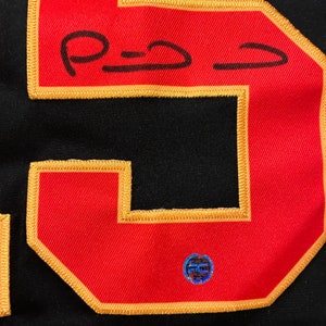 Puede incluir: Primer plano de una camiseta negra con un gran n&uacute;mero 5 rojo con contorno dorado. El n&uacute;mero tiene una firma con tinta negra. Una peque&ntilde;a pegatina hologr&aacute;fica redonda est&aacute; adherida a la camiseta.