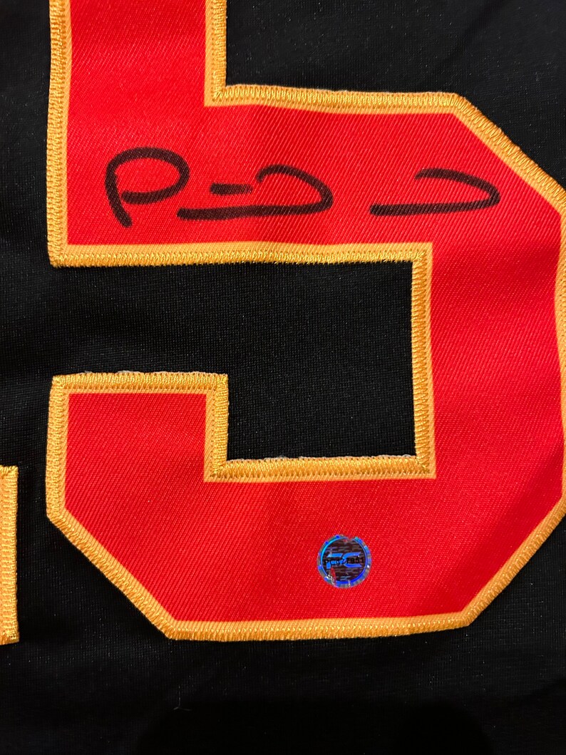 Puede incluir: Primer plano de una camiseta negra con un gran n&uacute;mero 5 rojo con contorno dorado. El n&uacute;mero tiene una firma con tinta negra. Una peque&ntilde;a pegatina hologr&aacute;fica redonda est&aacute; adherida a la camiseta.