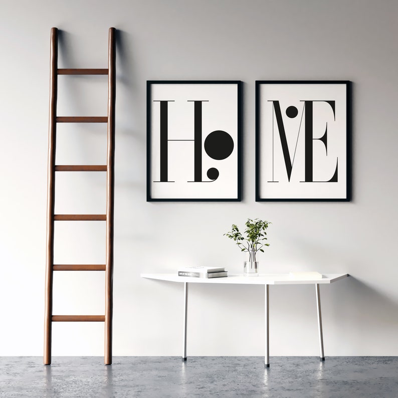 May include: Two framed art prints with the word "HOME" in a minimalist black and white design. A wooden ladder leans against the wall. A white table with a vase of greenery and books sits below the prints.